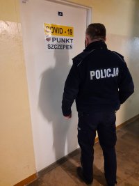 Umundurowany policjant oczekujący przed drzwiami punktu szczepień przeciwko Cowid-19