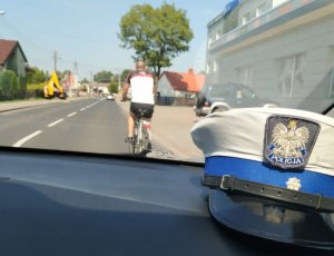 zdjęcie zrobione z radiowozu policyjnego Na kokpicie auta leży policyjna czapka , dalej widać drogę,  po której jedzie rowerzysta