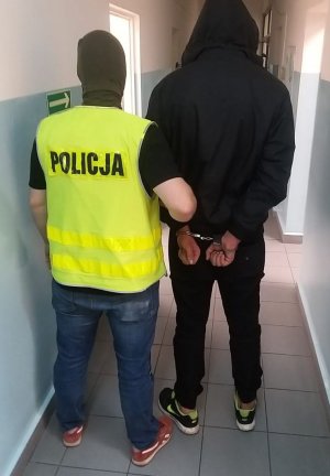 policjant służby kryminalnej ubrany po cywilnemu oraz w kamizelkę policyjną koloru żółtego z napisem policja prowadzi mężczyznę zatrzymanego, który ma założone kajdanki na ręce trzymane z tyłu. zdjęcie w korytarzu komendy.