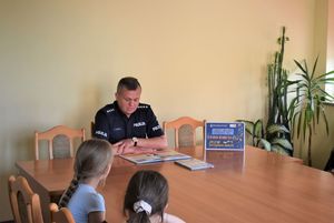 dzieci i policjant siedzą przy stole w pomieszczeniu komisariatu. policjant czyta książkę