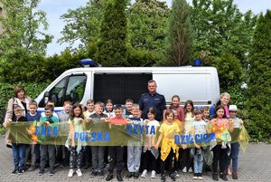 dzieci i policjant przy radiowozie . zdjęcie zrobione na placu wewnętrznym  komisariatu. dzieci trzymają napis  cała polska czyta dzieciom