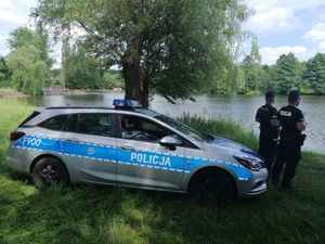 umundurowani policjanci nad zbiornikiem wodnym, radiowóz stoi przy wodzie w tle drzewa i woda