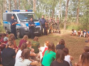 las, policjanci stoją przy radiowozie . prowadzą pogadankę z młodzieżą - harcerzami. Dzieci siedzą na ziemi. W tle rozbite namioty