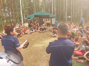 las, policjanci stoją przy radiowozie . prowadzą pogadankę z młodzieżą - harcerzami. Dzieci siedzą na ziemi. W tle rozbite namioty