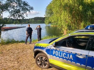 policjanci nad zbiornikiem wodnym, kontrolują bezpieczeństwo, przy jeziorze radiowóz.