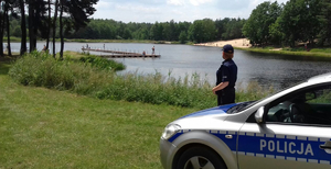 policjantka nad zbiornikiem wodnym, kontroluje bezpieczeństwo nad wodą, w tle pomost na którym chodzą wczasowicze, przy jeziorze radiowóz.