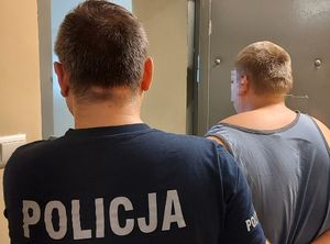 policjant prowadzi podejrzanego. zdjęcie zrobione w pomieszczeniu służbowym. na fotografii widać tych mężczyzna od głowy do pasa od tyłu.