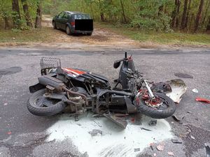 na zdjęciu widok uszkodzonego w wyniku wypadku motoroweru, który lezy na drodze. w tle droga , las i samochód osobowy.
