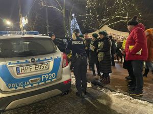 na świątecznym jarmarku policjantka rozdaje kamizelki odblaskowe. widoczny radiowóz policyjny dalej uczestnicy jarmarku.