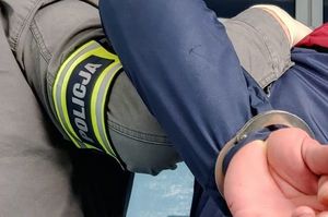 widok ramienia mężczyzny z opaska z napisem policja oraz widoczne są dłonie osoby , która ma założone kajdanki na ręce trzymane z tyłu.