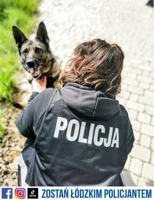 policyjny pies służbowy ze swoim przewodnikiem, pies ma założone szelki policyjne z napisem policja, przewodnik psa kuca przy nim ma założoną kamizelkę z napisem na plecach POLICA. Zdjęcie zrobione w plenerze. Pod zdjęciem napis o treści Zostań Łódzkim Policjantem.