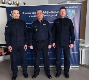komendant i dwóch policjantów, stoją przy ściance z napisem komenda powiatowa policji w Bełchatowie, zdjęcie zrobione w auli komendy.