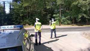 policjanci w czasie działań, stoją przy drodze i badają prędkość pojazdów, obok widać częściowo radiowóz policyjny, dalej drzewa.