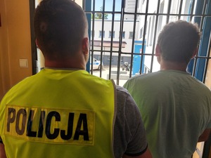 policjant prowadzi zatrzymanego. zdjęcie zrobione w pomieszczeniu służbowym. W tle zakratowane okno, przez które widać dziedziniec komendy i zaparkowany radiowóz policyjny