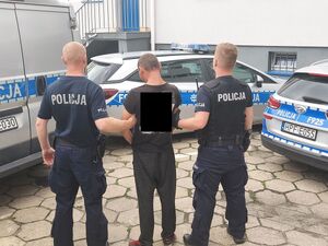 policjanci prowadza zatrzymanego. zdjęcie zrobione na placu wewnętrznym komendy. w tle radiowozy policyjne i część  budynku komendy
