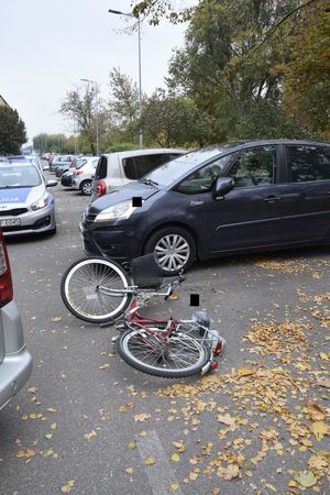 miejsce wypadku drogowego, potrącenie rowerzysty. rower leży na drodze  obok zaparkowane na parkingu auta, dalej radiowóz policyjny i droga osiedlowa