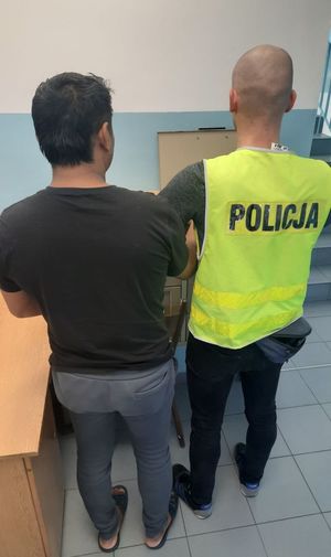 Zatrzymany mężczyzna stoi tyłem, obok funkcjonariusz w kamizelce z napisem policja.