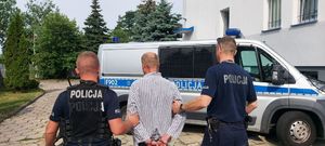 policjanci prowadzą podejrzanego, mężczyzna na ręce trzymane z tyłu ma założone kajdanki, dalej widać plac wewnętrzny prze komendą policji, budynek komendy w tylnej części oraz radiowóz.