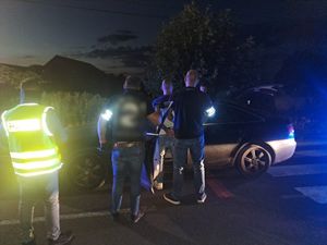 Zdjęcie wykonane nocą. Przy samochodzie tyłem stoi zatrzymany mężczyzna, po jego obu stronach policjanci w policyjnych opaskach na ramieniu. Pol lewej stronie funkcjonariusz w kamizelce z napisem policja.
