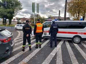 na zdjęciu trzech funkcjonariuszy: policji straży pożarnej i straży miejskiej stoją w rejonie drogi dbając o bezpieczeństwo, w tle radiowóz straży i ulica.