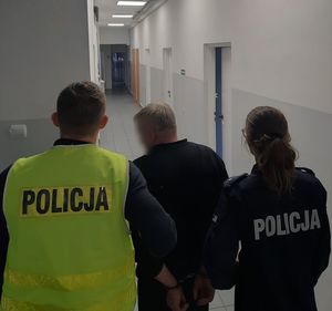 Policjanci prowadzą zatrzymanego mężczyznę, który ma założone kajdanki na ręce trzymane z tyłu.