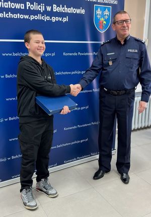 na zdjęciu policjant i chłopiec. komendant ściska dłoń chłopca, w tle baner z napisem komendant powiatowa policji w Bełchatowie.