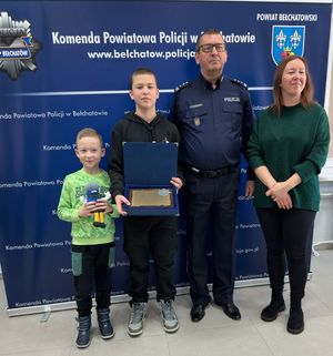 na zdjęciu policjant dwóch chłopców i kobieta. W tle baner z napisem komendant powiatowa policji w Bełchatowie.