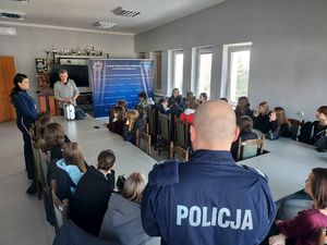 uczniowie w auli komendy, siedzą przy stolikach, policjant rozmawia z nimi o policyjnej służbie.