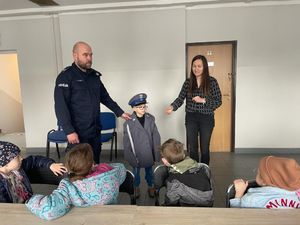 policjant rozmawia z dzieciakami o pracy policjanta, obok stoi chłopiec który ma założoną marynarkę i czapkę policyjną.
