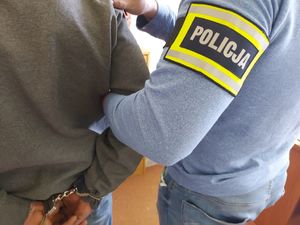 nieumundurowany policjant, który prowadzi zatrzymaną osobę. zdjęcie przedstawia przedramię policjanta na którym znajduje się opaska z napisem policja, widać też dłonie mężczyzny, na które założone są kajdanki.