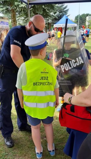 Pod policyjnym namiotem policjant z dziećmi uczestniczącymi w wydarzeniu Bezpieczne Wakacje w Powiatowy Centru Sportu, pokazuje im wyposażenie policjanta do służby. chłopiec ma założoną kamizelkę z napisem jedz bezpiecznie