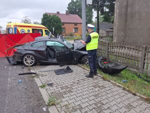 śmiertelny wypadek w miejscowości mierzynów. Na zdjęciu policjant na miejscu zdarzenia, wykonuje oględziny rozbitego samochodu osobowego, ktory przodem wbił się w latarnię uliczną, dalej czerwony parawan i karetka pogotowia.