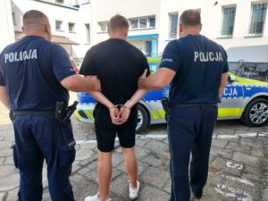 Dwóch policjantów prowadzi zatrzymanego mężczyznę. Zdjęcie zrobione na placu wewnętrzny komendy, widoczny radiowóz policyjny oraz budynek komendy policji.