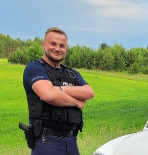 zdjęcie umundurowanego policjanta, dalej widoczna łąka i drzewa, narożnik pojazdu.