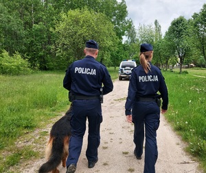 policyjny patrol skweru, dwóch umundurowanych policjantów z psem służbowym patroluje teren parku, w tle teren zielony dalej radiowóz.