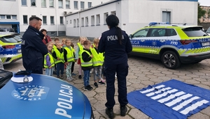 policjantka w czasie spotkania z przedszkolakami na dziedzińcu komendy Policji. Funkcjonariuszka prowadzi zajęcia z dziećmi ucząc ich zasad bezpiecznego przemieszczania się po drodze.