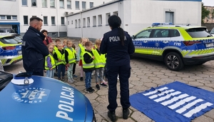 policjantka w czasie spotkania z przedszkolakami na dziedzińcu komendy Policji. Funkcjonariuszka prowadzi zajęcia z dziećmi ucząc ich zasad bezpiecznego przemieszczania się po drodze.