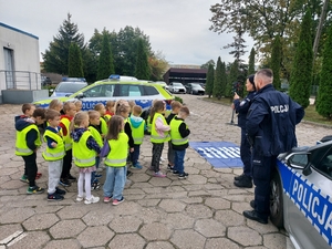 policjanci w czasie spotkania z przedszkolakami na dziedzińcu komendy Policji. Funkcjonariuszka prowadzi zajęcia z dziećmi ucząc ich zasad bezpiecznego przemieszczania się po drodze.