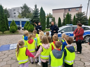 policjantka w czasie spotkania z przedszkolakami na dziedzińcu komendy Policji. Funkcjonariuszka prowadzi zajęcia z dziećmi ucząc ich zasad bezpiecznego przemieszczania się po drodze.
