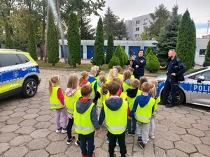 policjantka w czasie spotkania z przedszkolakami na dziedzińcu komendy Policji. Funkcjonariuszka prowadzi zajęcia z dziećmi ucząc ich zasad bezpiecznego przemieszczania się po drodze.
