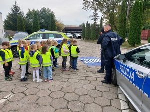 policjantka w czasie spotkania z przedszkolakami na dziedzińcu komendy Policji. Funkcjonariuszka prowadzi zajęcia z dziećmi ucząc ich zasad bezpiecznego przemieszczania się po drodze.