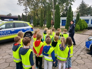 policjantka w czasie spotkania z przedszkolakami na dziedzińcu komendy Policji. Funkcjonariuszka prowadzi zajęcia z dziećmi ucząc ich zasad bezpiecznego przemieszczania się po drodze.