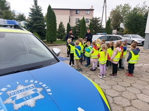 policjantka w czasie spotkania z przedszkolakami na dziedzińcu komendy Policji. Funkcjonariuszka prowadzi zajęcia z dziećmi ucząc ich zasad bezpiecznego przemieszczania się po drodze.