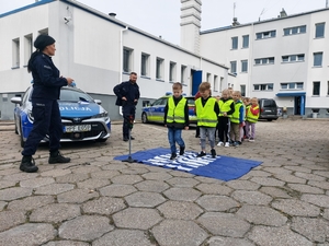 policjantka w czasie spotkania z przedszkolakami na dziedzińcu komendy Policji. Funkcjonariuszka prowadzi zajęcia z dziećmi ucząc ich zasad bezpiecznego przemieszczania się po drodze.