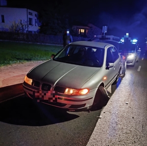 samochód osobowy z uszkodzonym przednim lewym kołem stoi na drodze, za nim widoczny radiowóz policyjny.