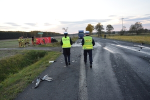 miejsce śmiertelnego wypadku drogowego, dwóch umundurowanych policjantów na drodze, dalej pojazd uczestnicząc w zdarzeniu oraz czerwony parawan zabezpieczający miejsce wypadku.