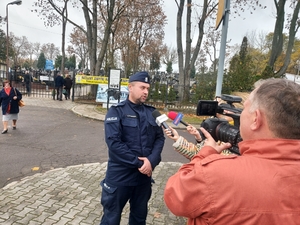 umundurowany policjant udziela wywiadu mediom, w tle cmentarz.