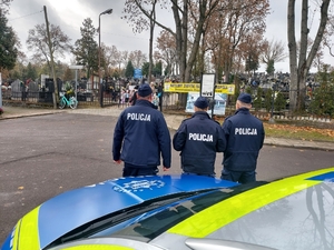 trzech policjantów stoi przy radiowozie, dbają o bezpieczeństwo w rejonie cmentarzy.