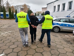 dwóch nieumundurowanych policjantów prowadzi zatrzymanego mężczyznę. zdjęcie zrobione na placu wewnętrznym komendy, w tle widoczne radiowozy policji.