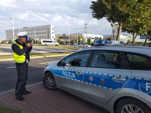 policjant stoi przy drodze, trzyma urządzenie do badania prędkości pojazdów, obok radiowóz, dalej droga i zabudowania.